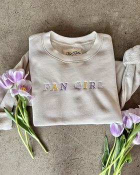 close up picture of a tan crewneck that says Fan Girl and there are flowers laying on the crew