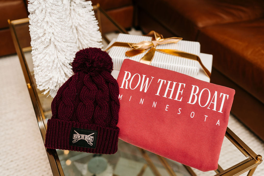 close up picture of a maroon winter hat with a Row the Boat detail and a maroon sweatshirt with Row the Boat screenprinted detail next to a Christmas trees and presents