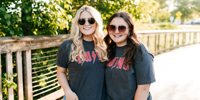 two girls on a bridge with trees in the background wearing the same faded gray t-shirt and sunglasses 