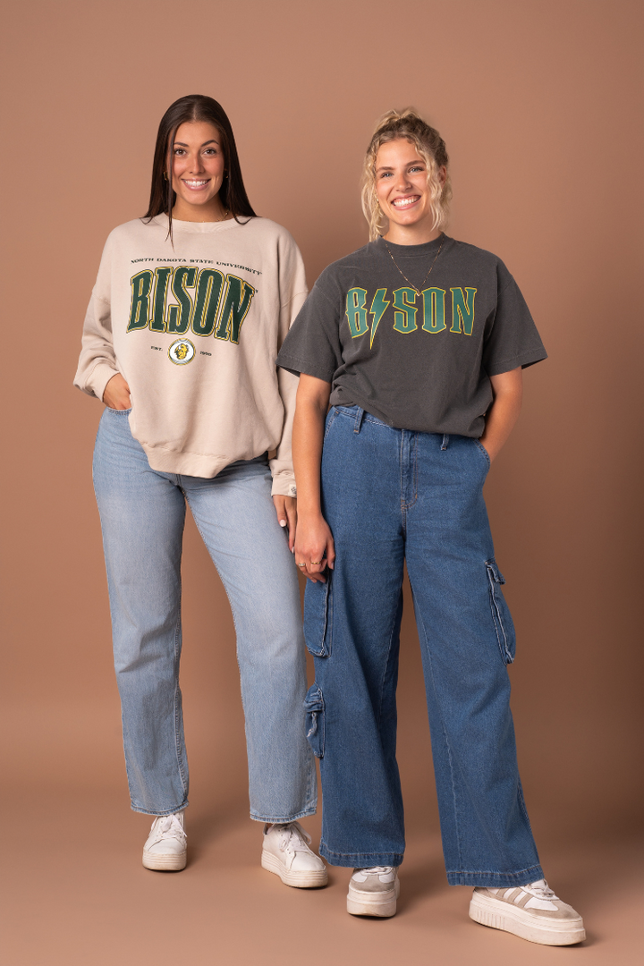 two girls wearing jeans and one has a NDSU Bison tan crewneck one and the other has a faded black Bison band tee on