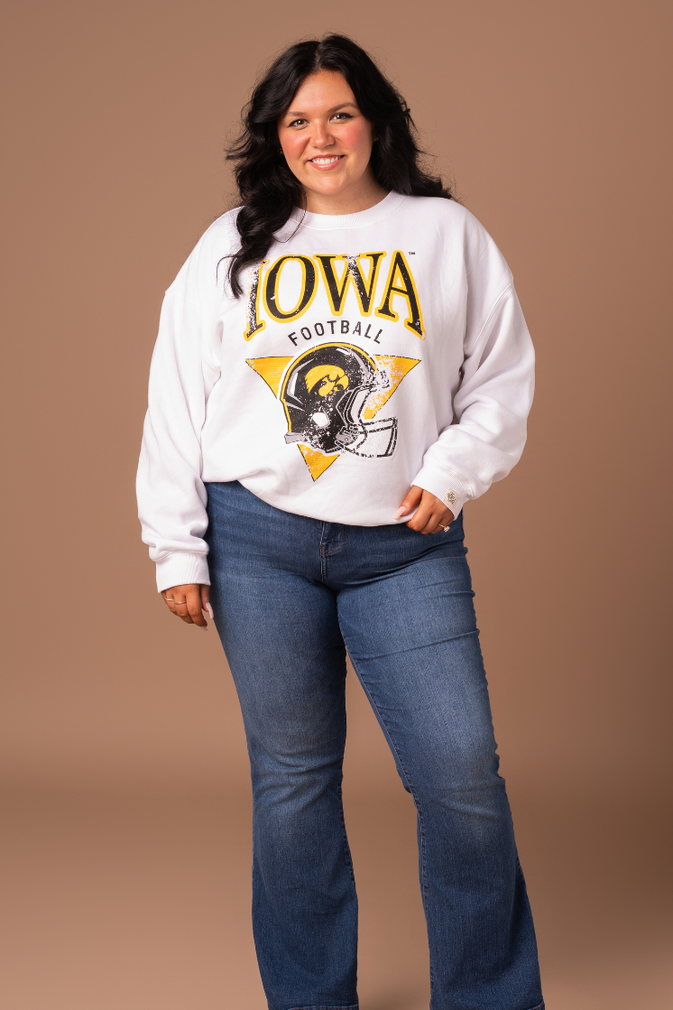 farther away photo of a woman wearing a women's fit, white crew neck sweatshirt. The sweatshirt has black and yellow screenprinted Iowa Hawkeyes football design with a helmet.