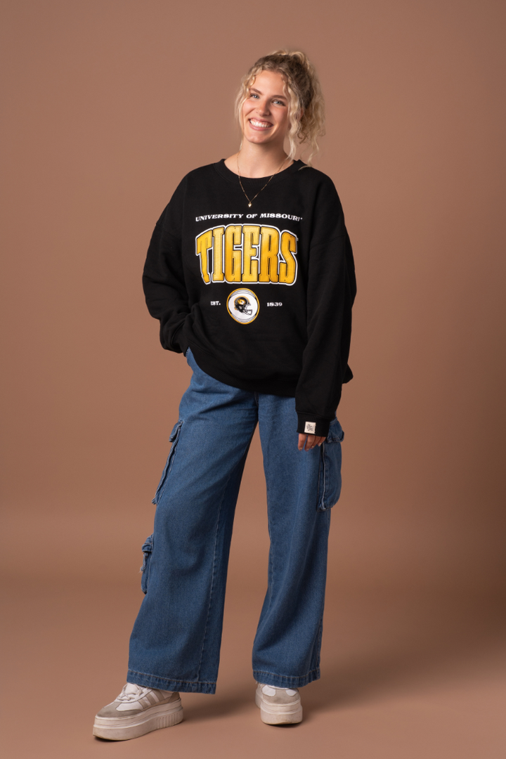 A full body photo of a girl wearing a black University of Missouri Tigers oversized crew neck with the established date and a football helmet in the graphic. She wearing this with jeans and sneakers. 