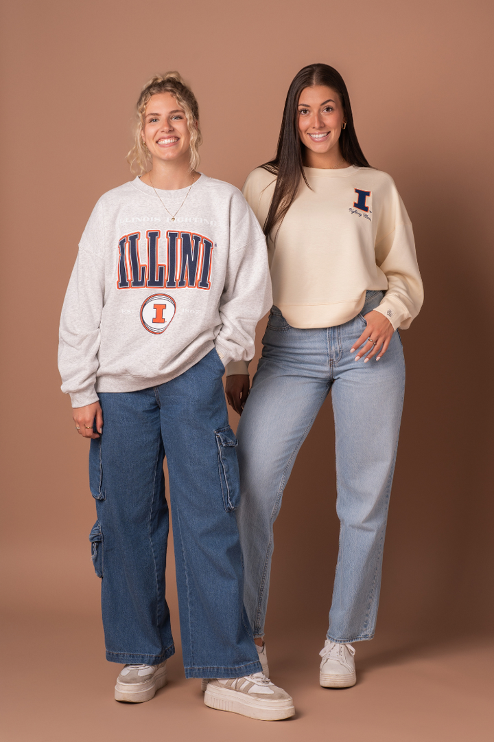 Group photo of two girls. One in athleisure type ivory crew neck sweater.With an embroidered University of Illinois logo with the cursive Fighting Illini text below it. The other girl is wearing more of a heavier cotton ash gray crew neck with a different, vintage "fighting Illini" design.