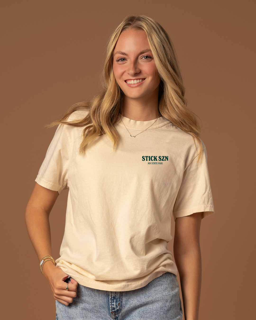 Girl wearing a beige t-shirt with dark green 'stick szn' and "mn state fair text. 