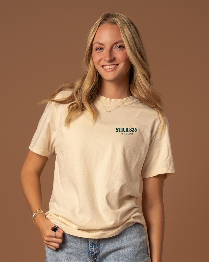 Girl wearing a beige t-shirt with dark green 'stick szn' and "mn state fair text. 