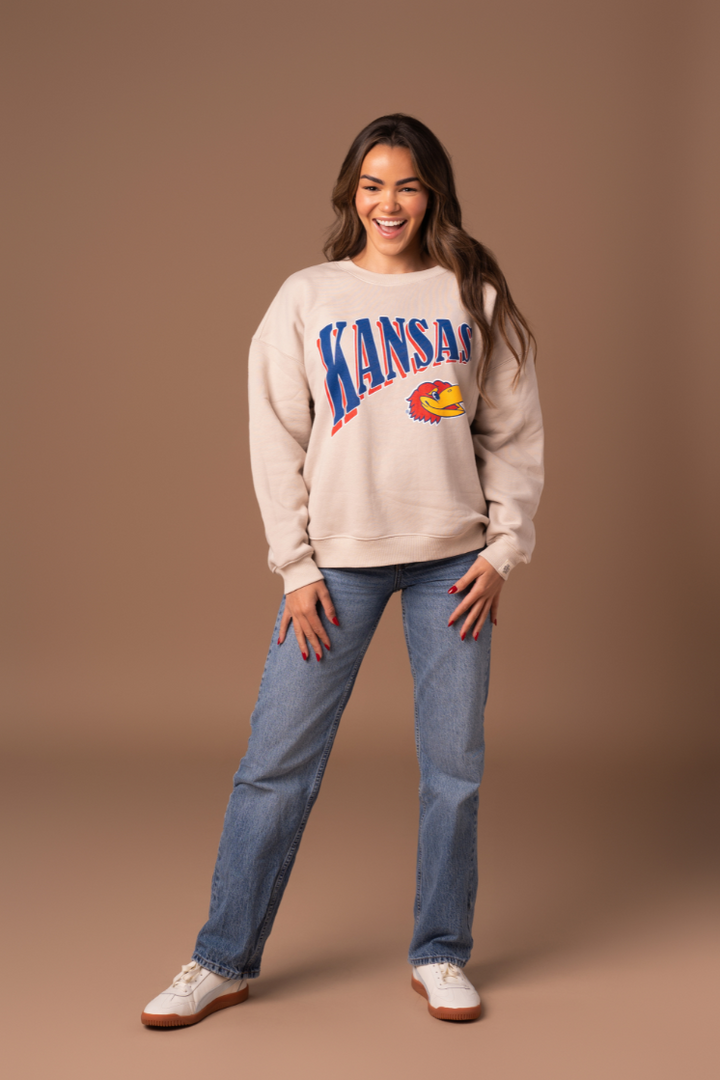 a full body image of a girl wearing jeans and a tan crewneck with Kansas screenprinted across the chest in blue and red outline with a Jayhawk logo below it