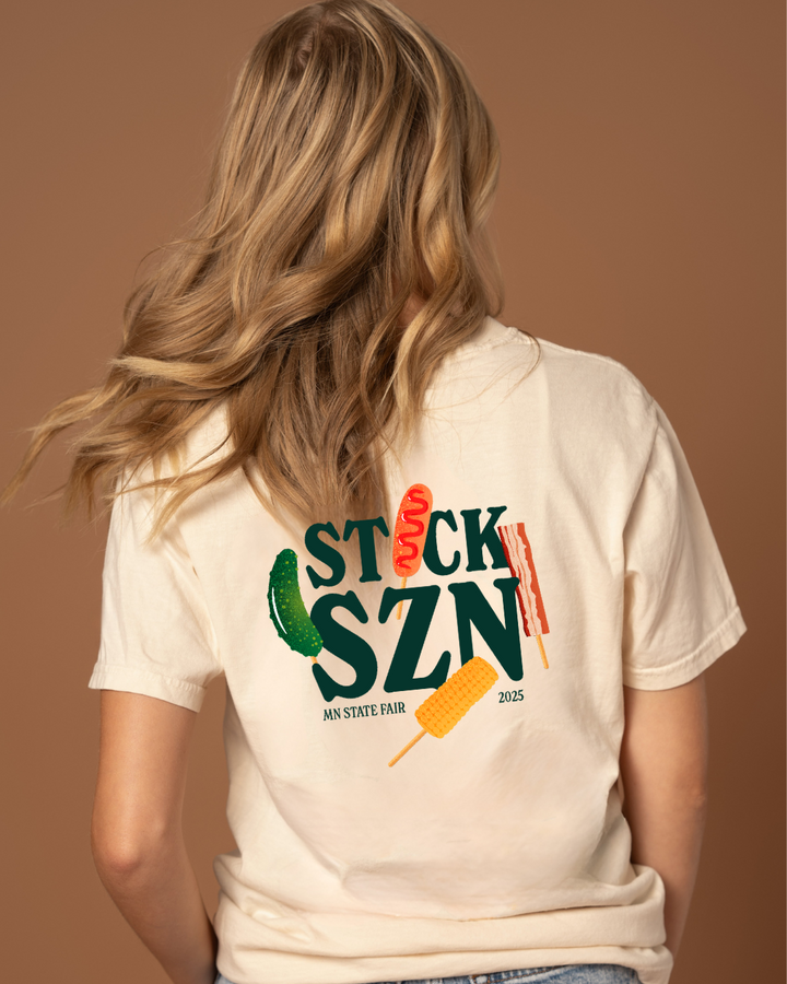 Girl wearing a beige t-shirt with dark green 'stick szn' text and colorful food on-a-stick graphics. 