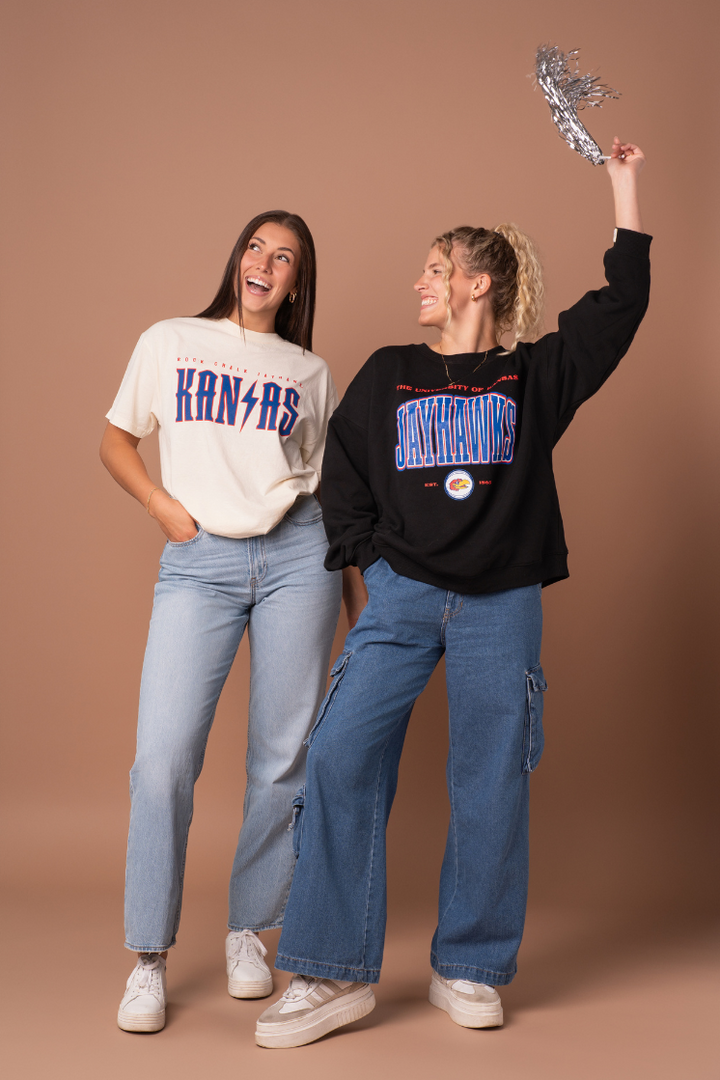 image of 2 girls posing and cheering while wearing two different Kansas styles, one cream t-shirt and one black crewneck