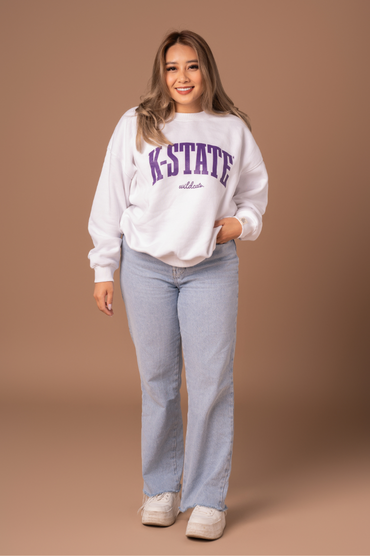 A full body photo of a girl wearing an oversized fleece lined white crew neck with a large purple K-State Wildcats design on the front. 