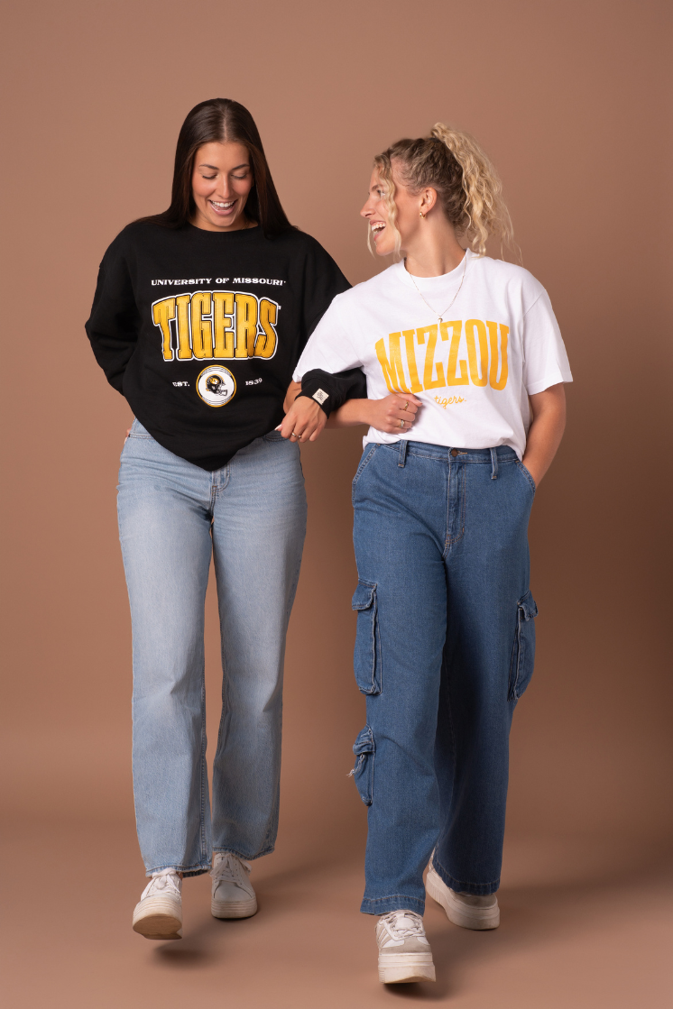 A photo of two girls linking arms a wearing Mizzou Tigers crew neck and t-shirt with blue jeans and sneakers. 