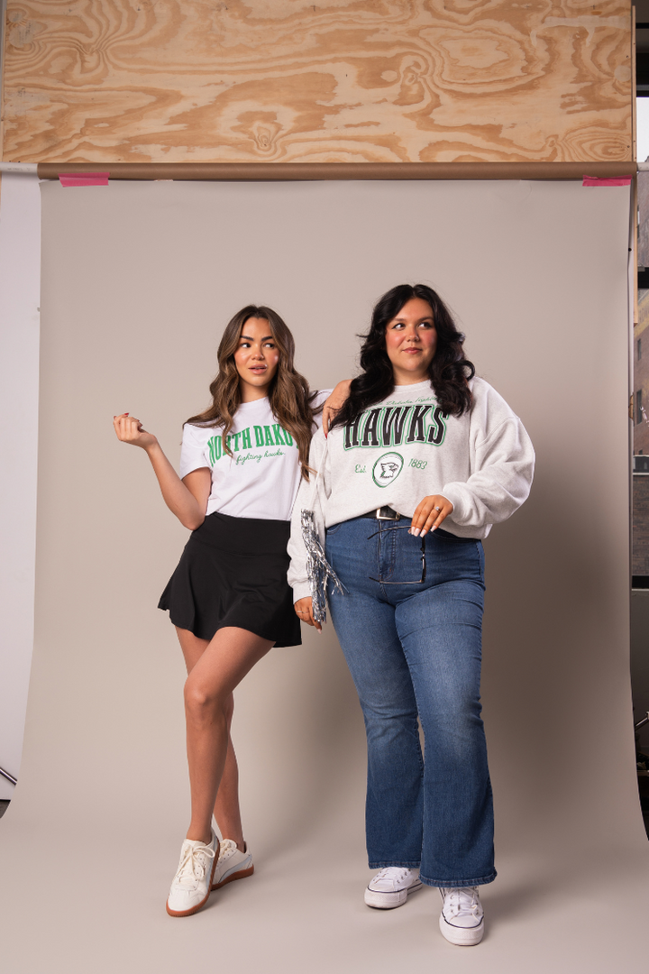 2 girls posing together, one in jeans and a gray crewneck with Hawks written across the chest and the other in a black skirt and white tshirt with North Dakota written in green