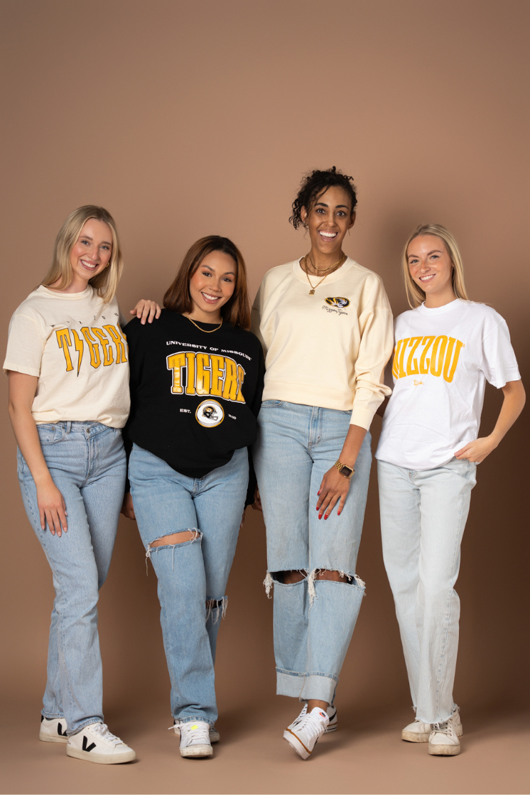 A photo of four girls wearing University of Missouri tops including a t-shirt, crew neck, and Supersoft crew neck. They all have different Mizzou Tigers designs on the front of the top. They are wearing these with jeans and sneakers. 