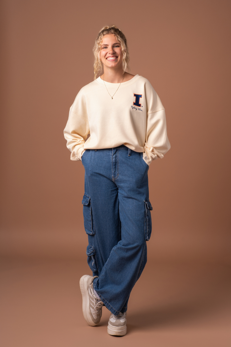 A woman with her hands in her pockets wearing a very soft, athleisure type ivory crew neck sweatshirt with an embroidered University of Illinois logo and cursive "fighting Illini" text below it. 