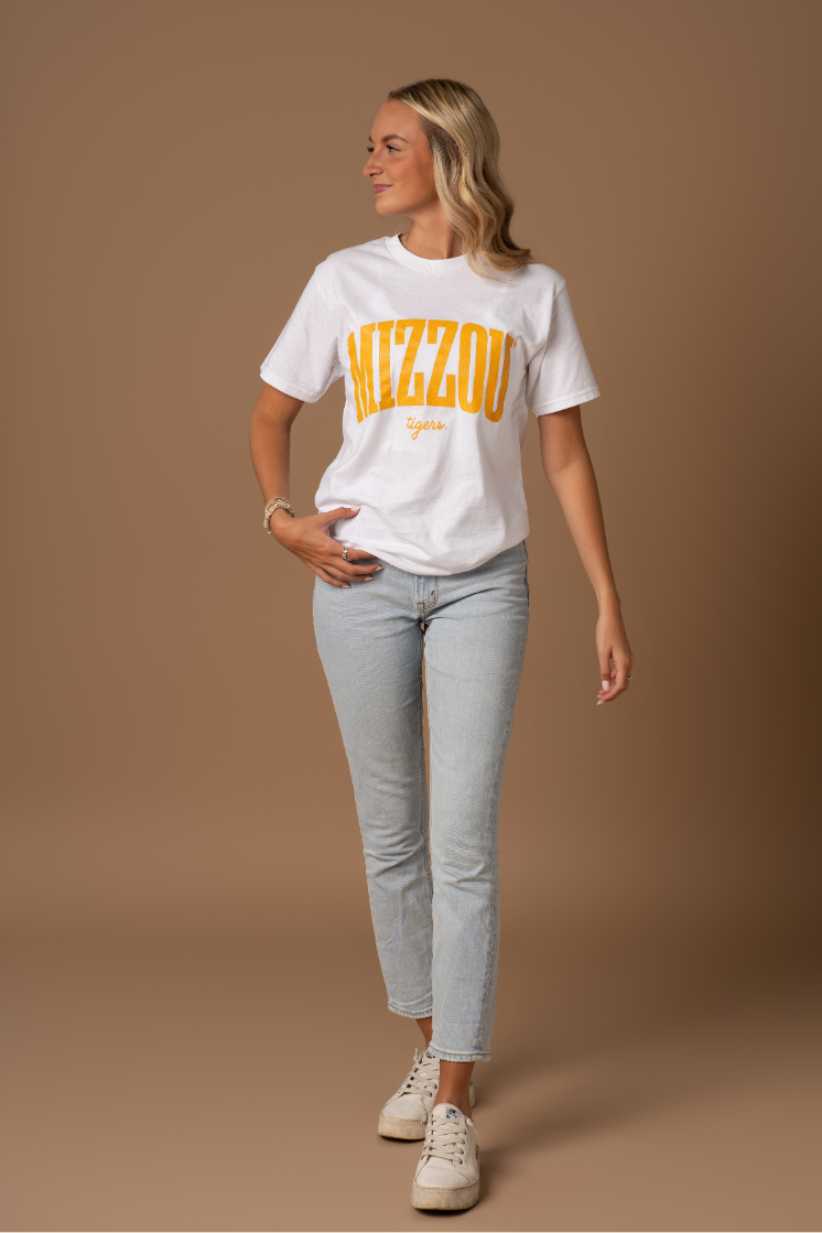 A full body photo of a girl wearing a white t-shirt with a Mizzou Tigers design on it in gold. She is wearing this with blue jeans and sneakers. 