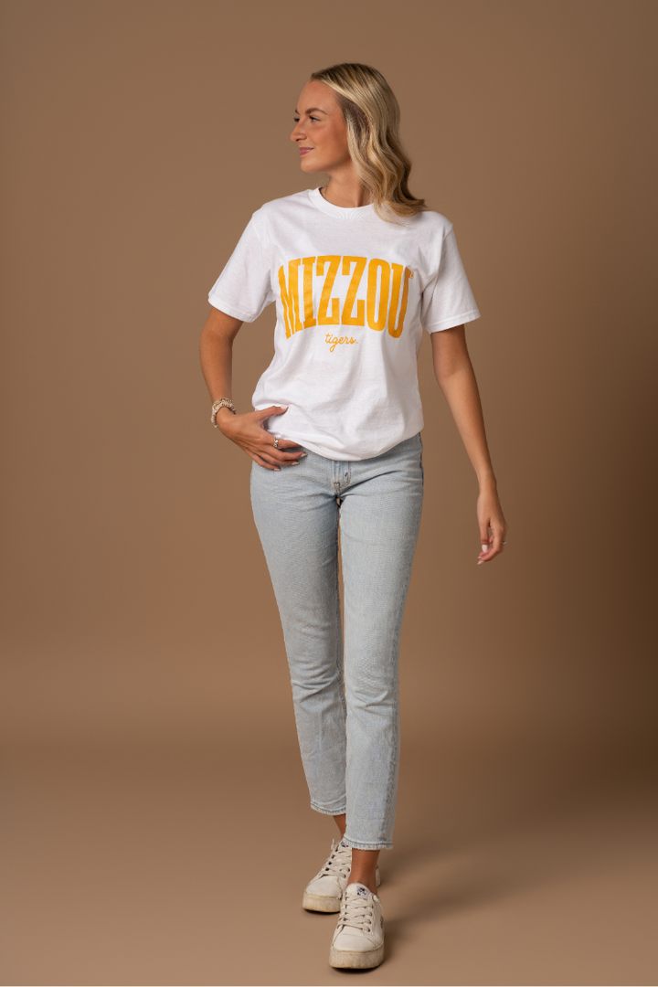 A full body photo of a girl wearing a white t-shirt with a Mizzou Tigers design on it in gold. She is wearing this with blue jeans and sneakers. 