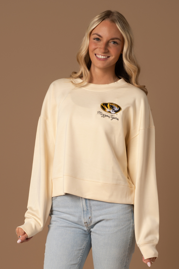 A close up photo of girl wearing a bone colored crew neck with an embroidered Mizzou Tigers logo and text on the upper chest. 