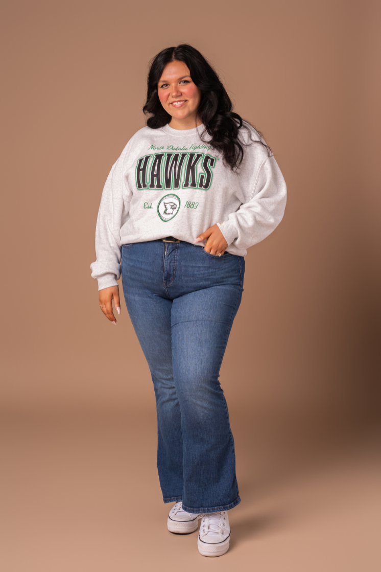 a full body image of a girl wearing jeans and a gray crewneck that says North Dakota Fighting Hawks and a hawk logo in green, black and gray decoration