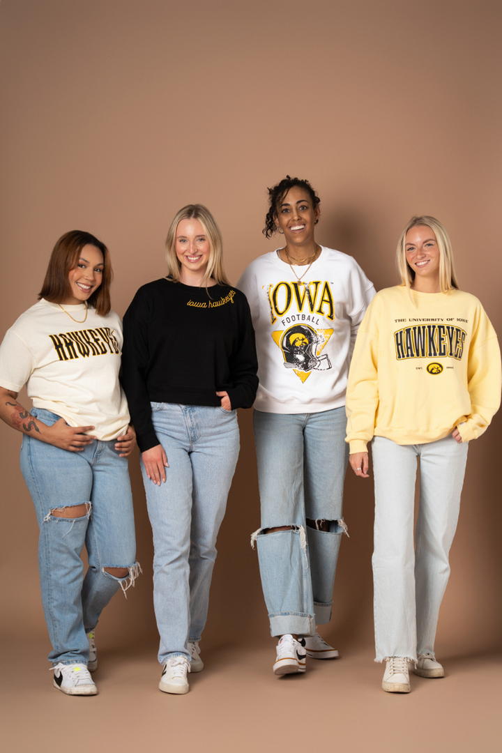 a group photo of four women wearing licensed university of iowa hawkeyes fan apparel. the apparel is crew neck sweatshirts and tshirts and are trendy, understated, and soft.