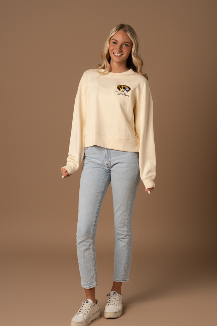 A full body photo of a girl wearing a crew neck with a Mizzou Tigers design on the left upper chest and blue jeans with sneakers. 