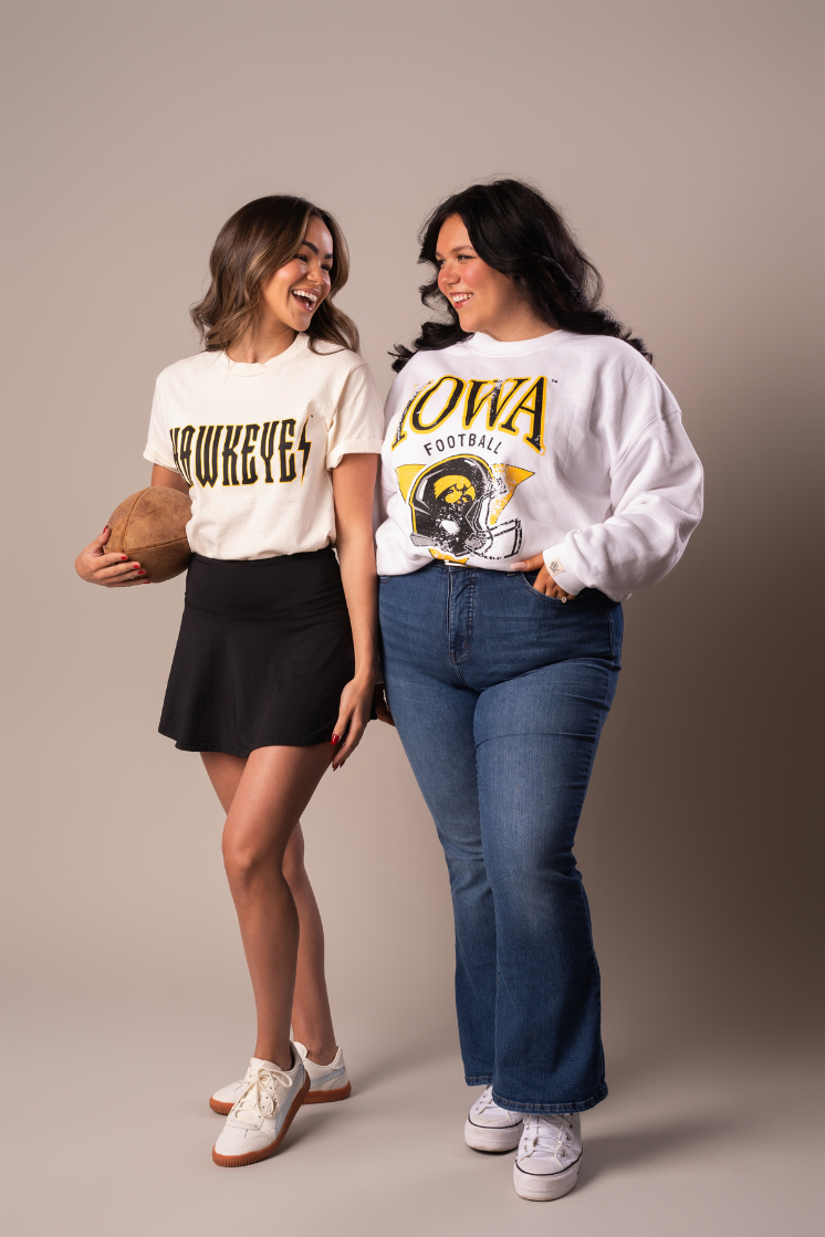 Two girls walking and flaunting their outfit. One wearing an ivory tshirt that has a trademarked Iowa Hawkeyes screenprinted design on it that resembles a trendy band tee. The other in a cotton crew neck sweatshirt with an iowa hawkeyes football design.