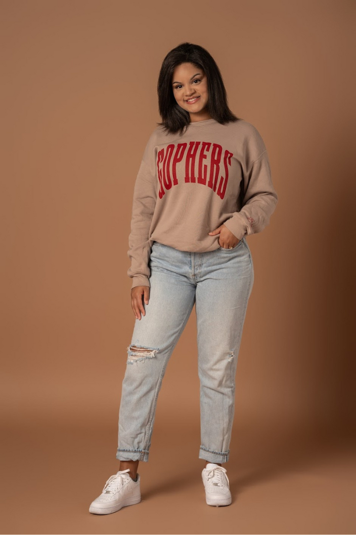 full body image of a girl wearing a tan colored crewneck with Gophers screenprinted maroon in big warped letters across the chest