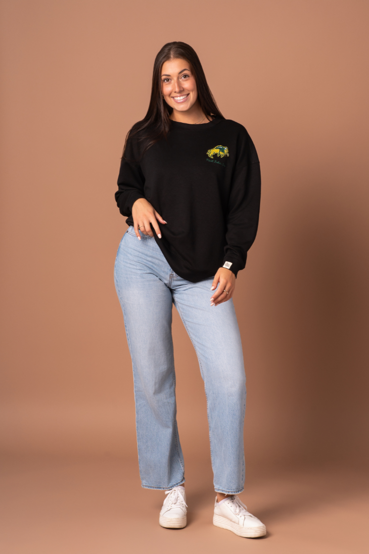 image of a girl standing and smiling with jeans and a black crewneck on, the crewneck has an embroidered NDSU Bison logo in the left pocketside