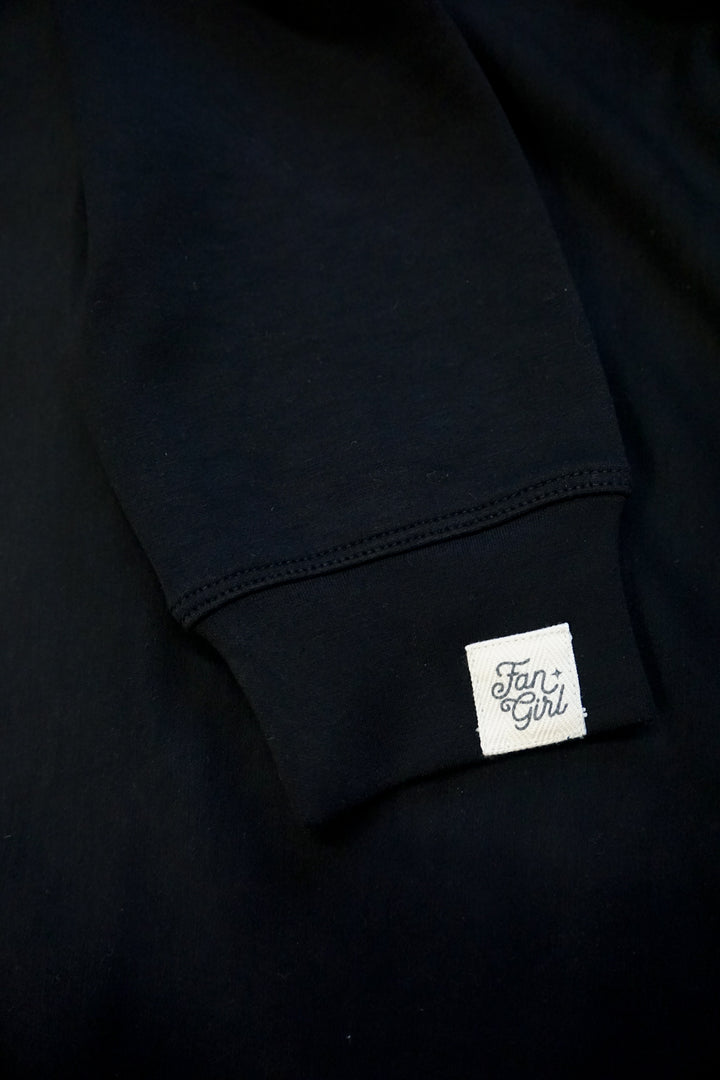 close up picture of a sleeve of a supersoft black crew neck sweatshirt with a small tan Fan Girl logo patch with black writing 