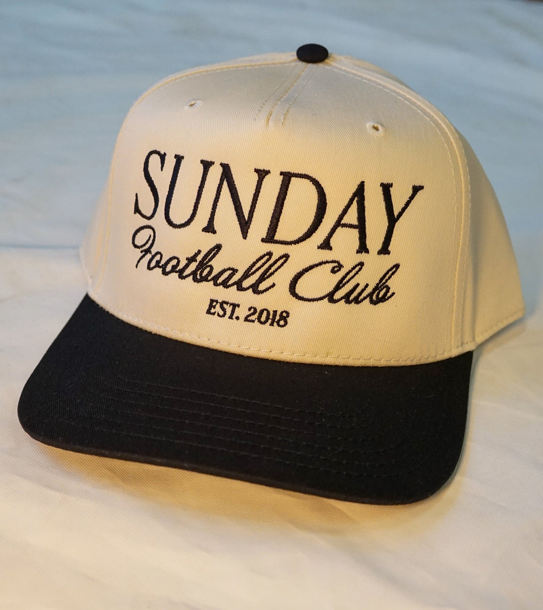 A close up of the front of a trucker hat. The brim of the hat is black and the head of the hat is a natural tan color with black premium embroidery that says "Sunday Football Club"