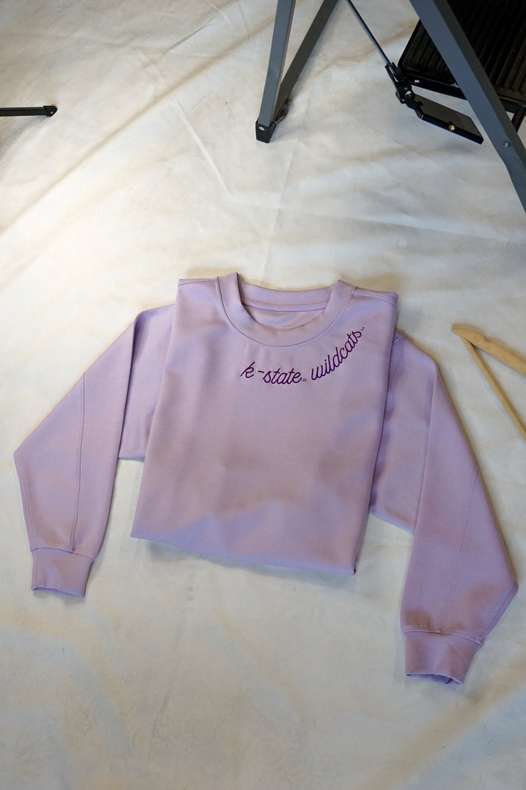 A staged flat-lay photo of a women's lavender crew beck with K-State Wildcats embroidery along the neck line. 