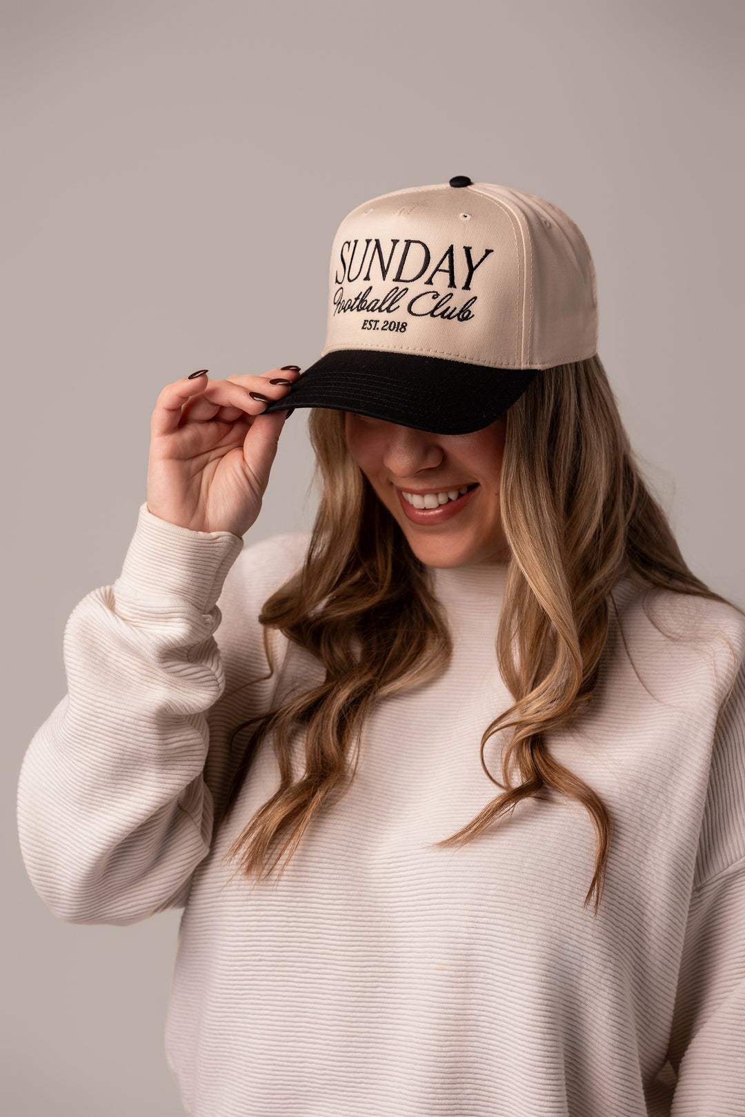 A girl wearing a trendy game day hat that has embroidered design on it that say s "Sunday Football Club". She's holding the black brim of the hat while the head of the hat is a natural tan color with black embroidery.