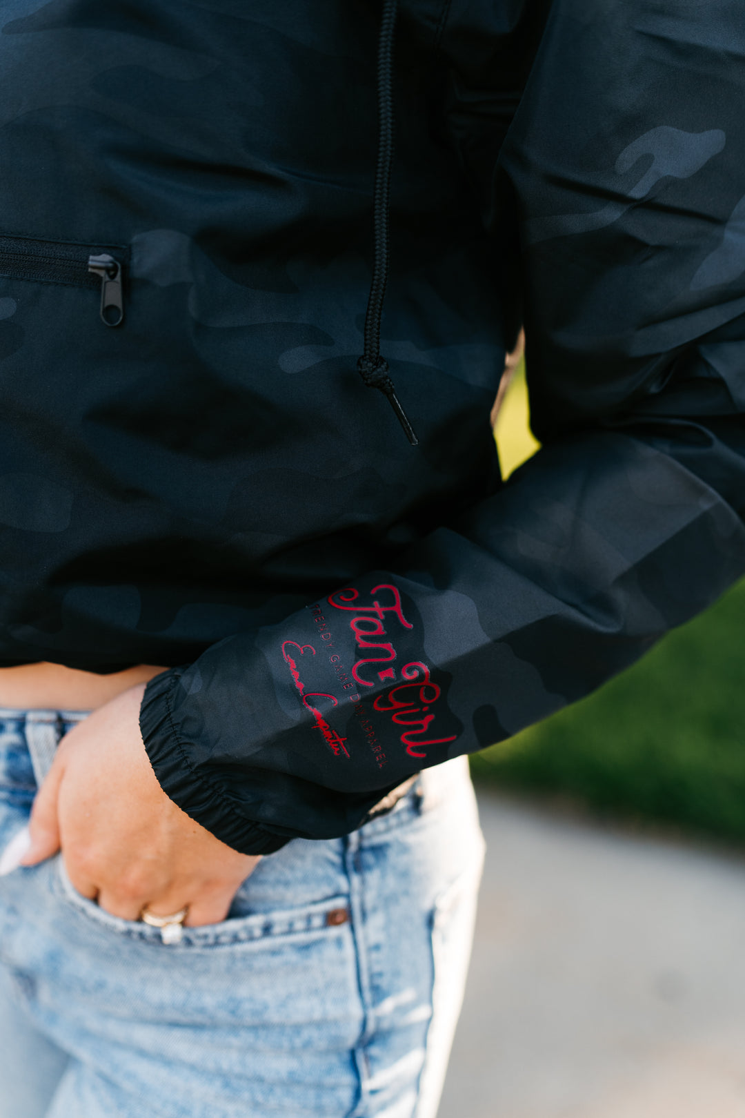 close up image of a sleeve of a camo windbreaker with a maroon Fan Girl logo and Emma Carpenter's signature