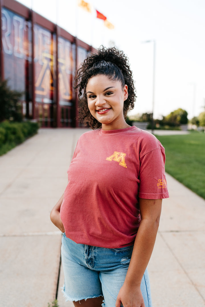 close up image of girl outside the Gophers hockey arena wearing a crimson colored t-shirt with a gold M screenprinted on the left pocketside of the tee