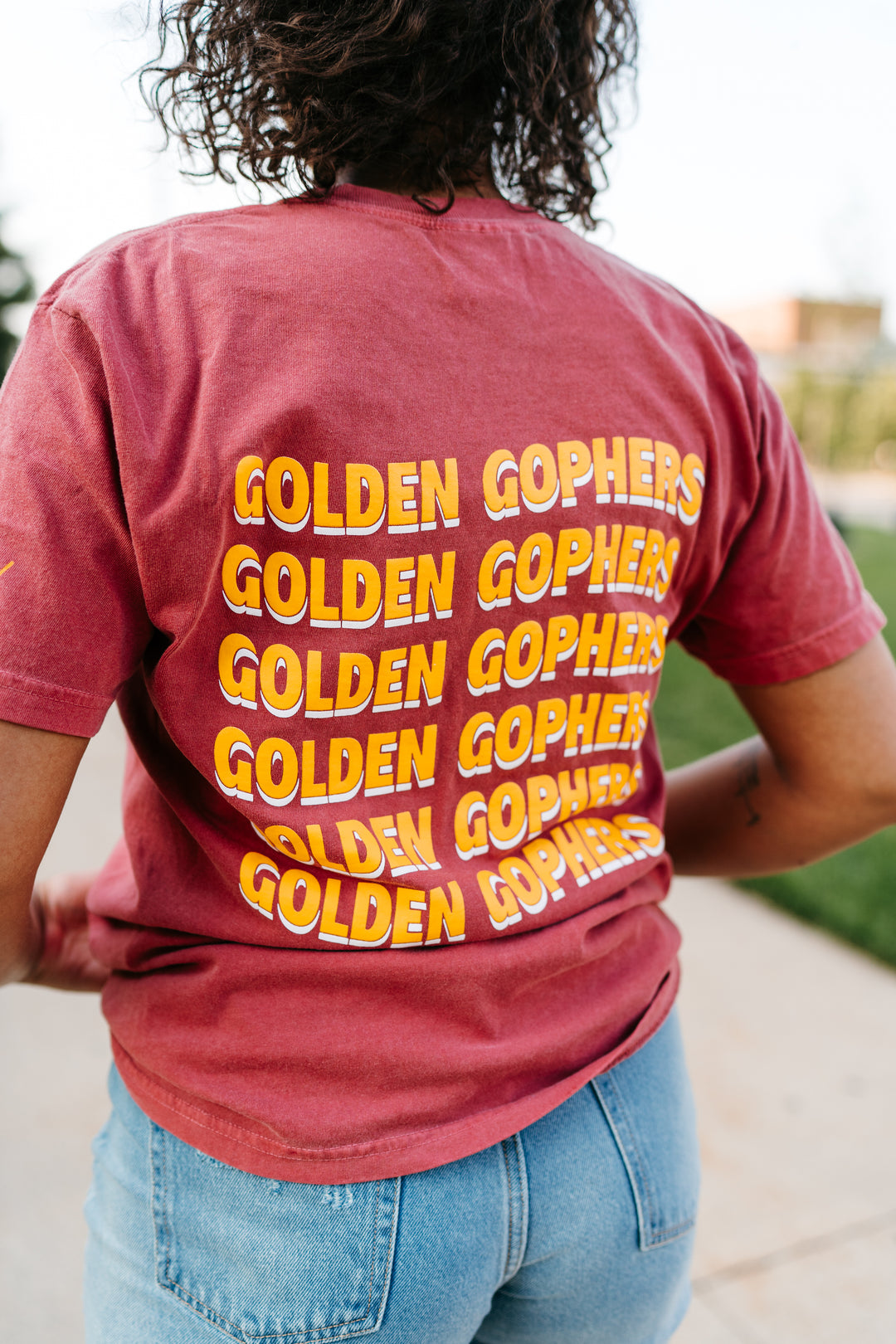 close up image of the back of girl outside wearing a crimson colored t-shirt with Golden Gophers screenprinted in gold that is stacked repeating six times