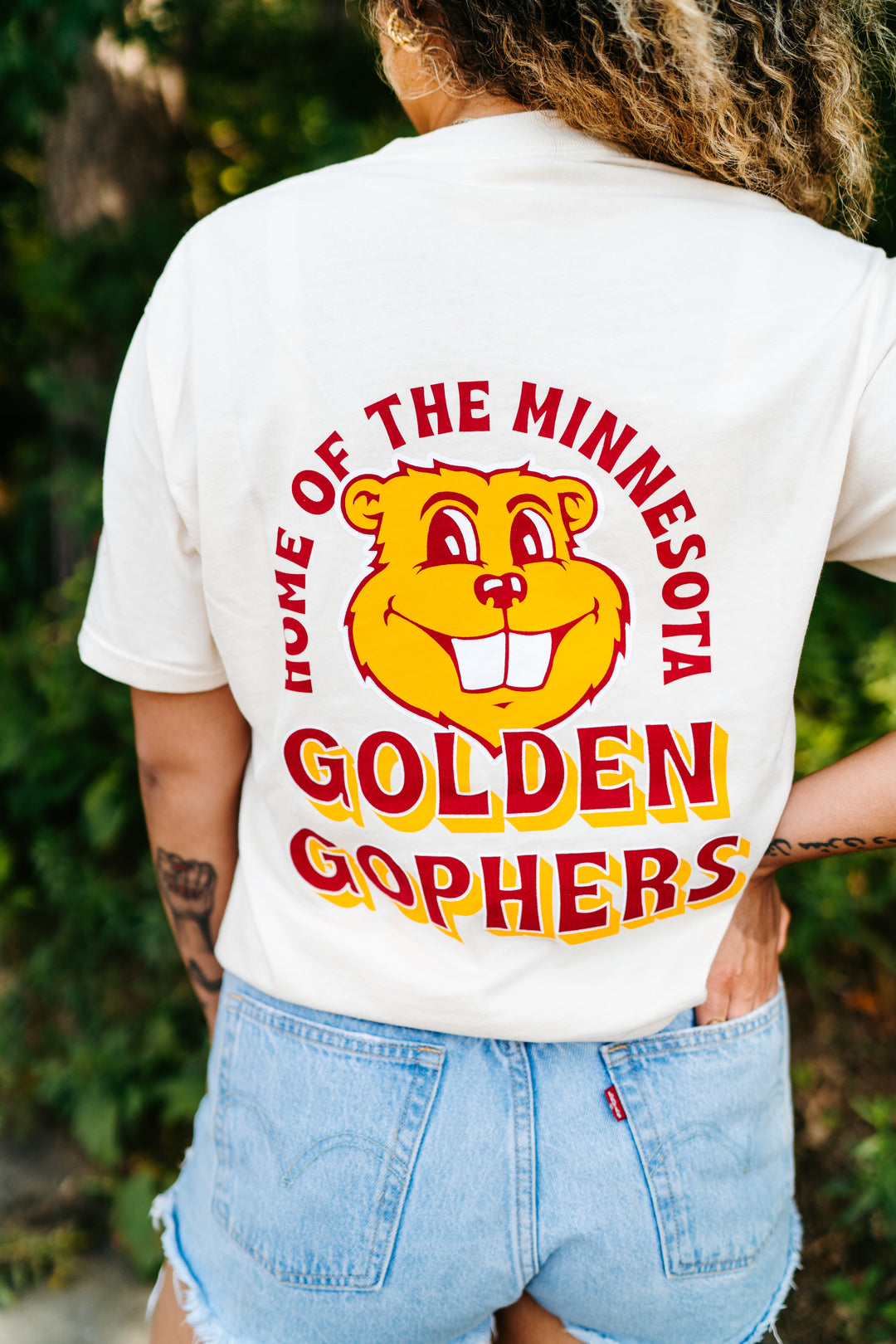 close up image of the back of a girl wearing a cream colored t-shirt with a Goldy graphic screenprinted in the center with Home of the Minnesota Golden Gophers outline in maroon and gold 