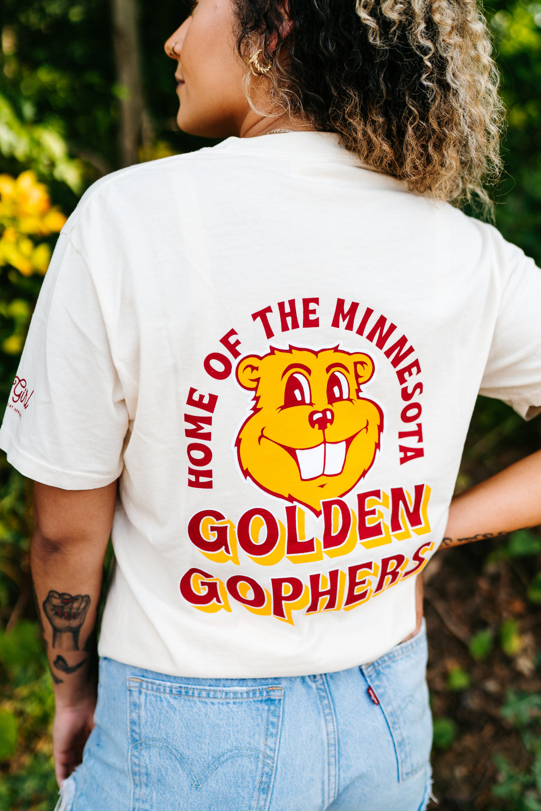 close up image of the back of a girl wearing a cream colored t-shirt with a Goldy graphic screenprinted in the center with Home of the Minnesota Golden Gophers outline in maroon and gold 