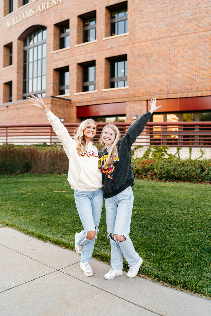 image of 2 girls with their hands in the air standing outside of Williams Arena and one is wearing an ivory Gophers hoodie and the other is wearing a black colored football crewneck