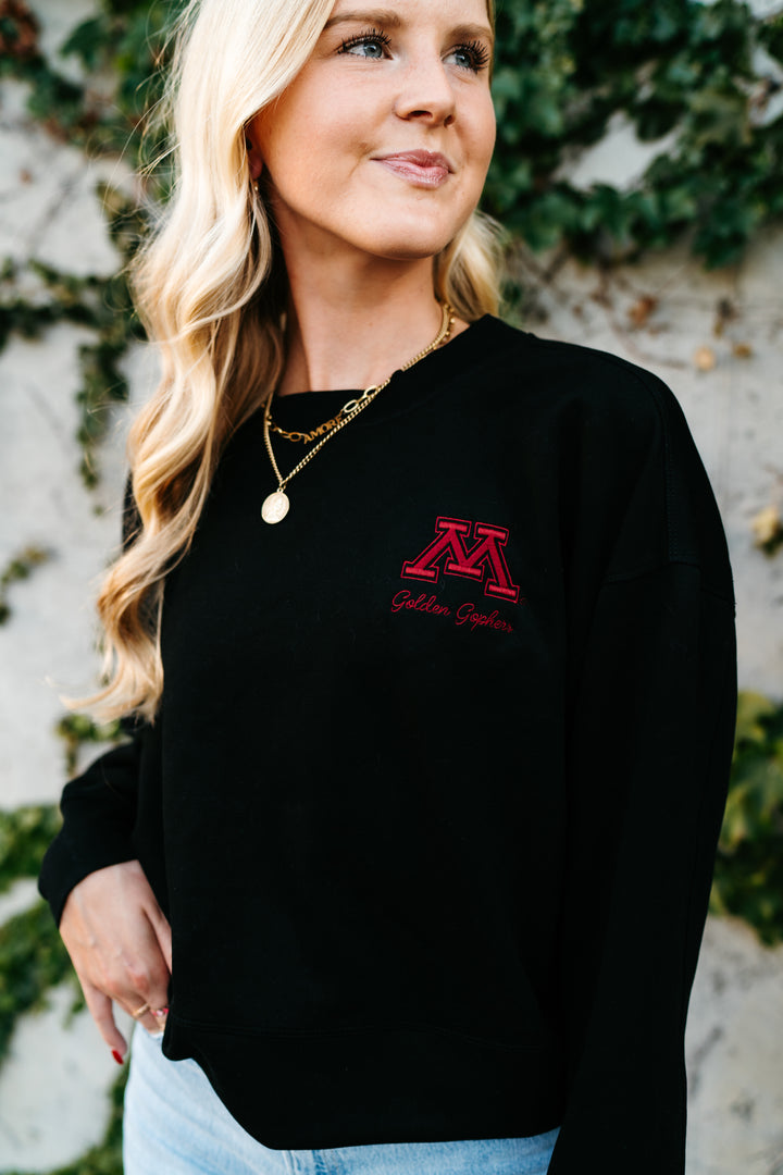 close up image of a girl outside wearing a black supersoft crewneck with a M logo and Golden Gophers embroidered in maroon on the left side pocket of the crew 