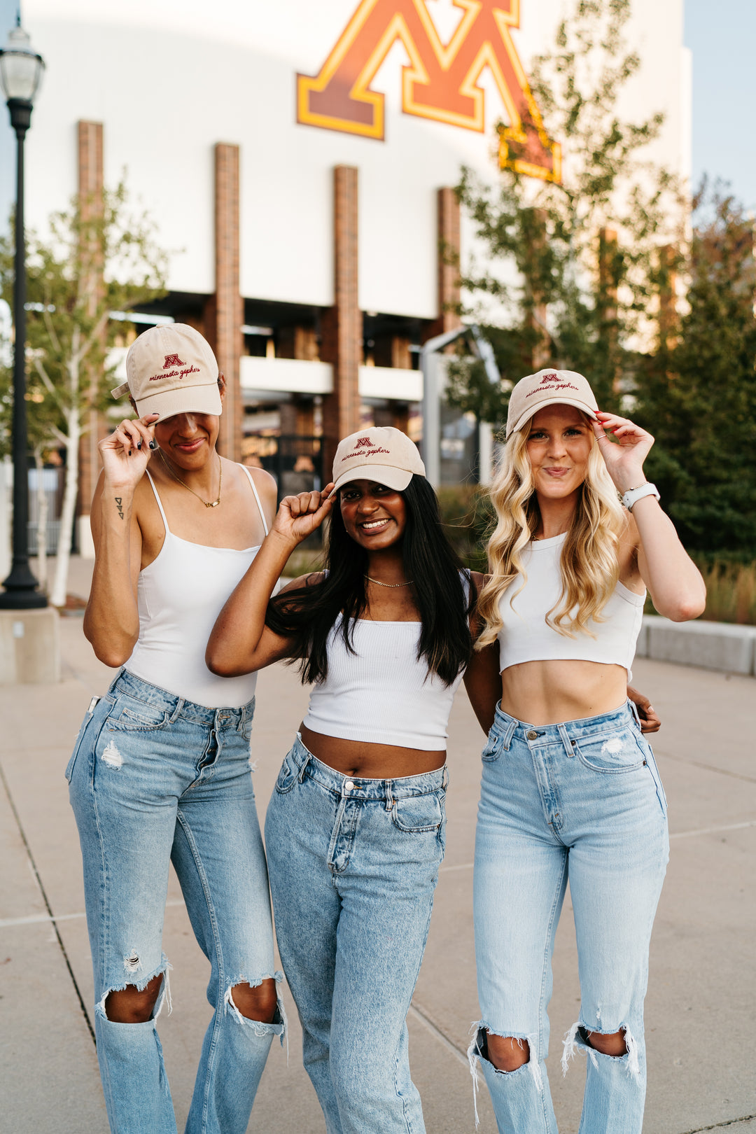 image of 3 girls outside wearing jeans, white tops and a tan hat with an M and minnesota gophers embroidered in cursive in maroon in front of a UMN building