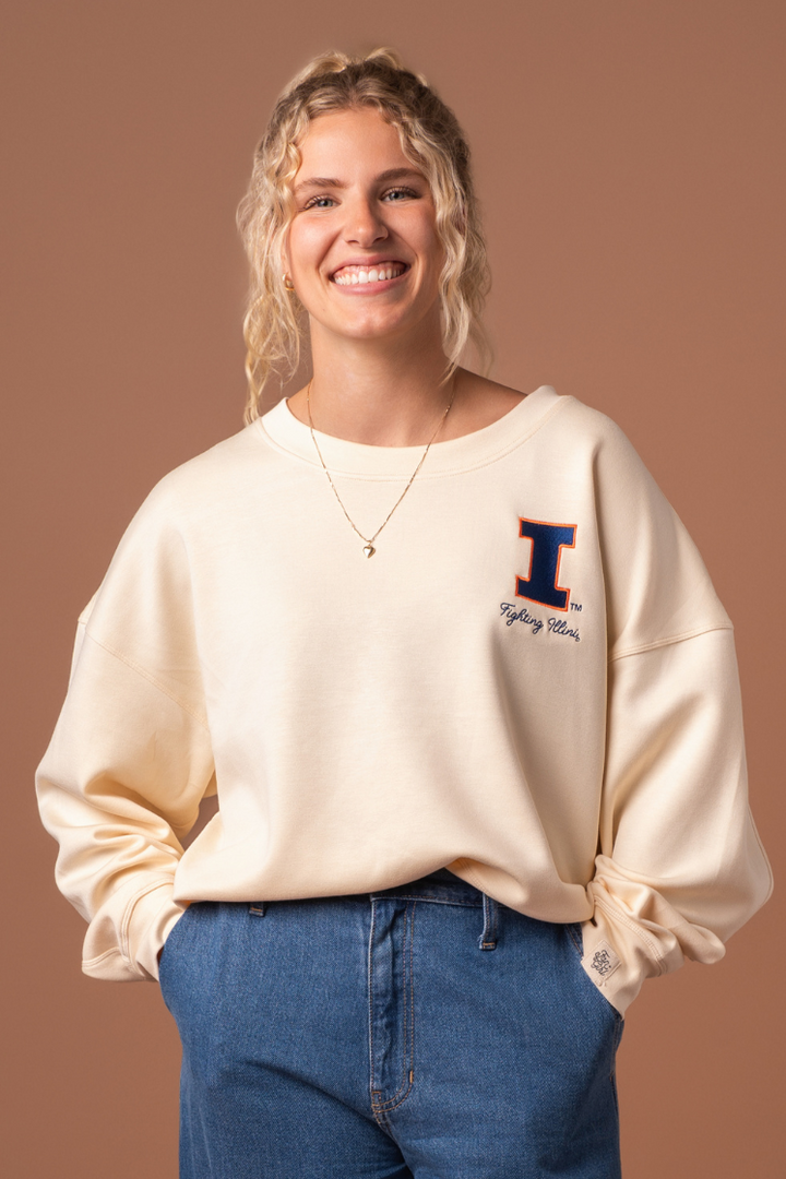 Close up of a woman wearing a very soft, athleisure type ivory crew neck sweatshirt with an embroidered University of Illinois logo and cursive "fighting Illini" text below it. 