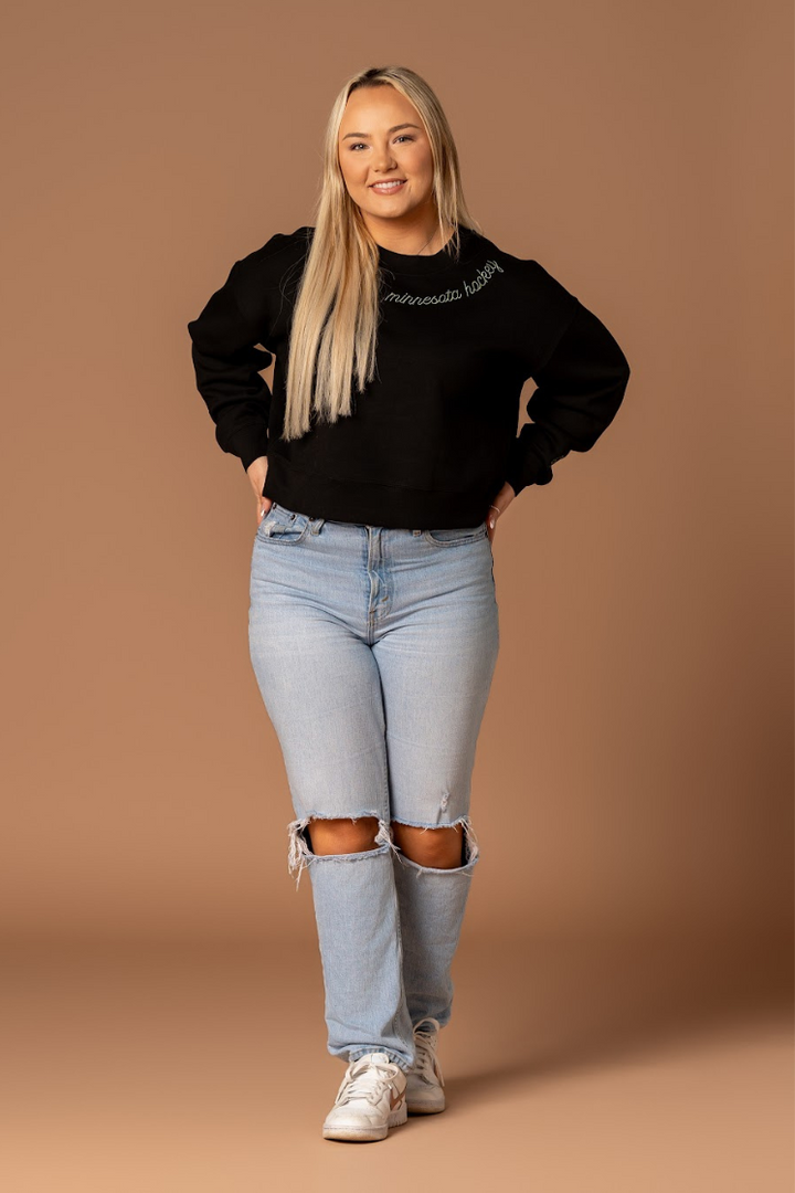 a farther away shot of a woman standing in an embroidered very soft black crew neck athleisure sweatshirt that says minnesota hockey