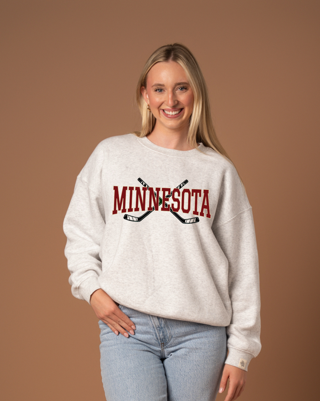 woman in a heather gray crew neck sweatshirt with maroon "minnesota" text and two green hockey sticks