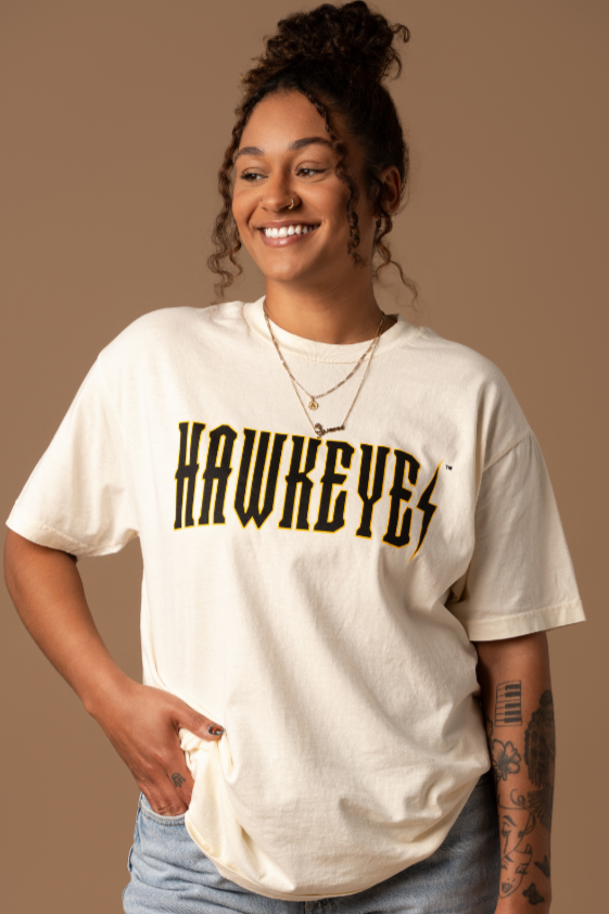 A close up of a woman wearing an ivory tshirt that has a trademarked Iowa Hawkeyes screenprinted design on it that resembles a trendy band tee.