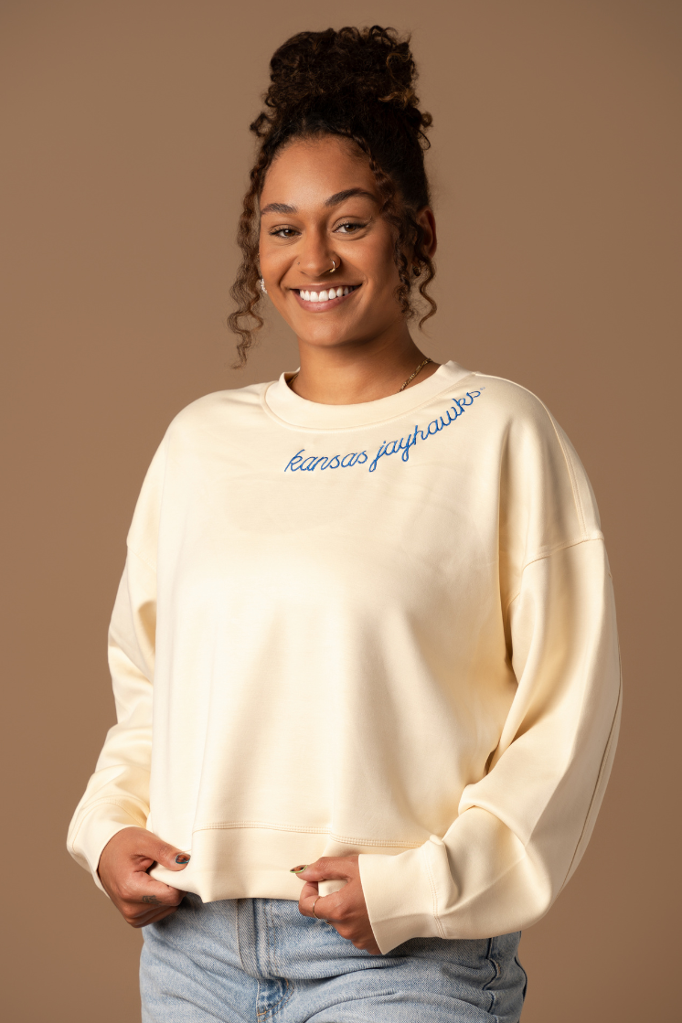 close up image of girl wearing a cream colored supersoft crew neck with kansas jayhawks embroidered around the collar in blue