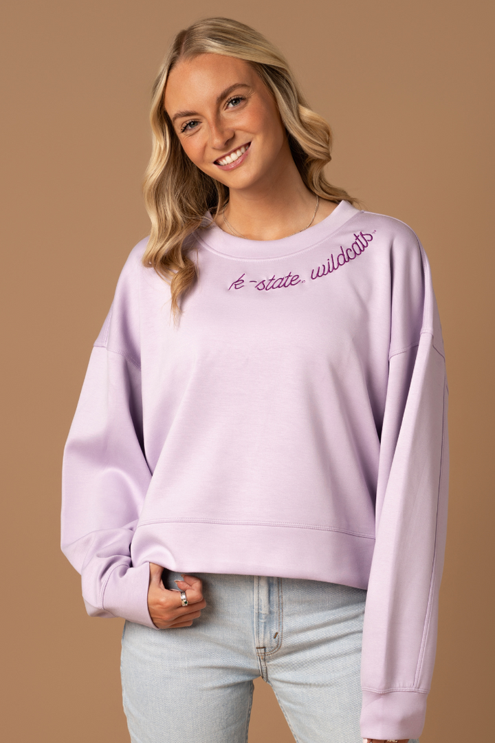 Close up image of a girl wearing a lavender crew neck with an embroidered K-State Wildcats design on the neckline.