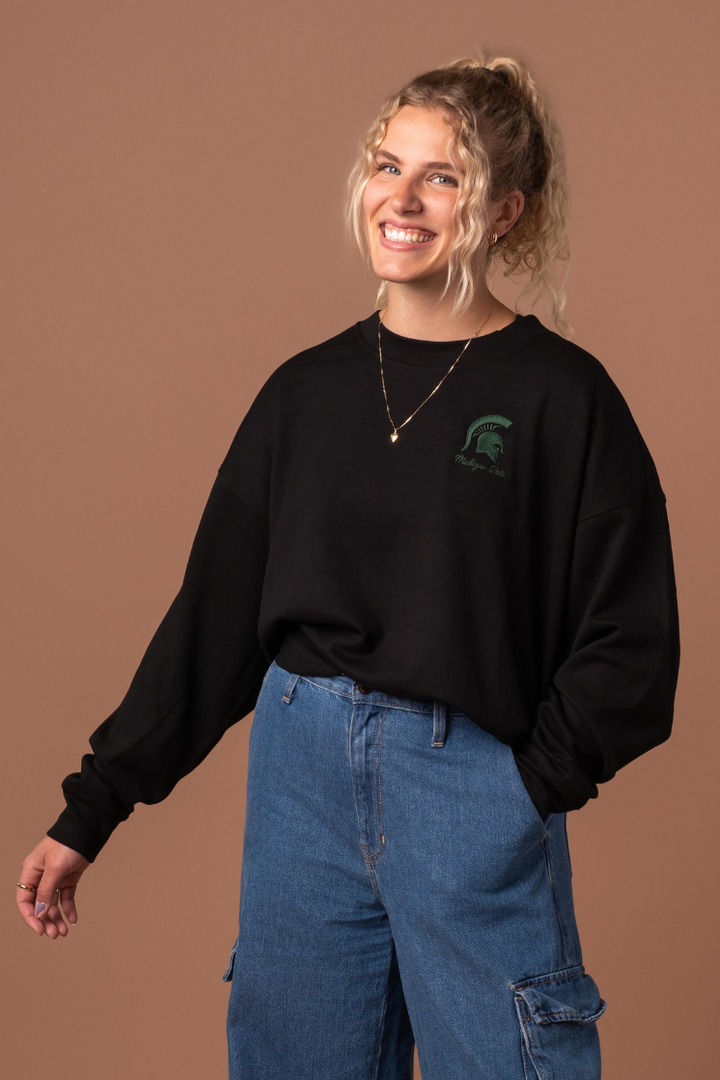 close up picture of a girl wearing a black Michigan State Supersoft Waist Length Crew Neck with a green embroidered  Spartan logo 