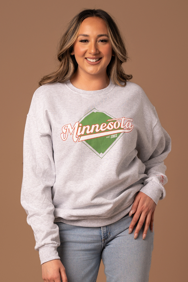 close up picture of a girl wearing a gray fleece lined crew neck with a screenprinted baseball field design in green with minnesota written in red and white cursive 