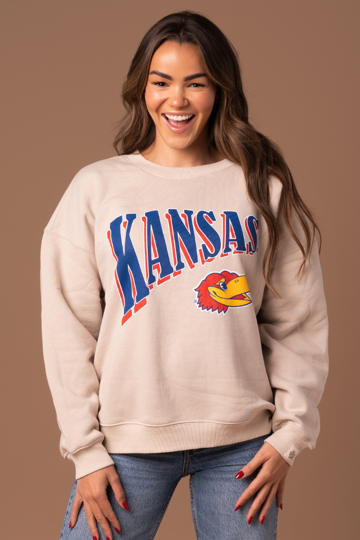 close up image of a girl wearing a tan crewneck with Kansas written across the chest with a Jayhawk graphic below it in red, blue and gold colors