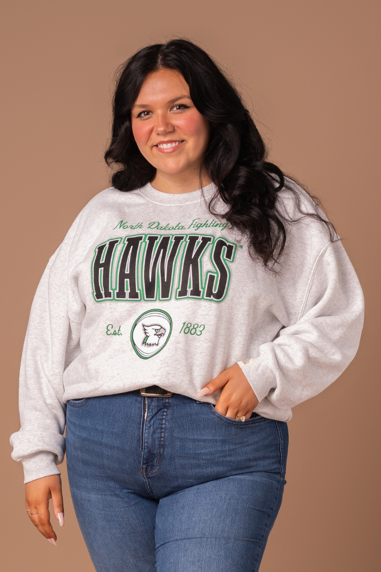 close up of a girl with her hand in her pocket and is wearing an ash gray crewneck with North Dakota Fighting Hawks written out in green, black, gray and white