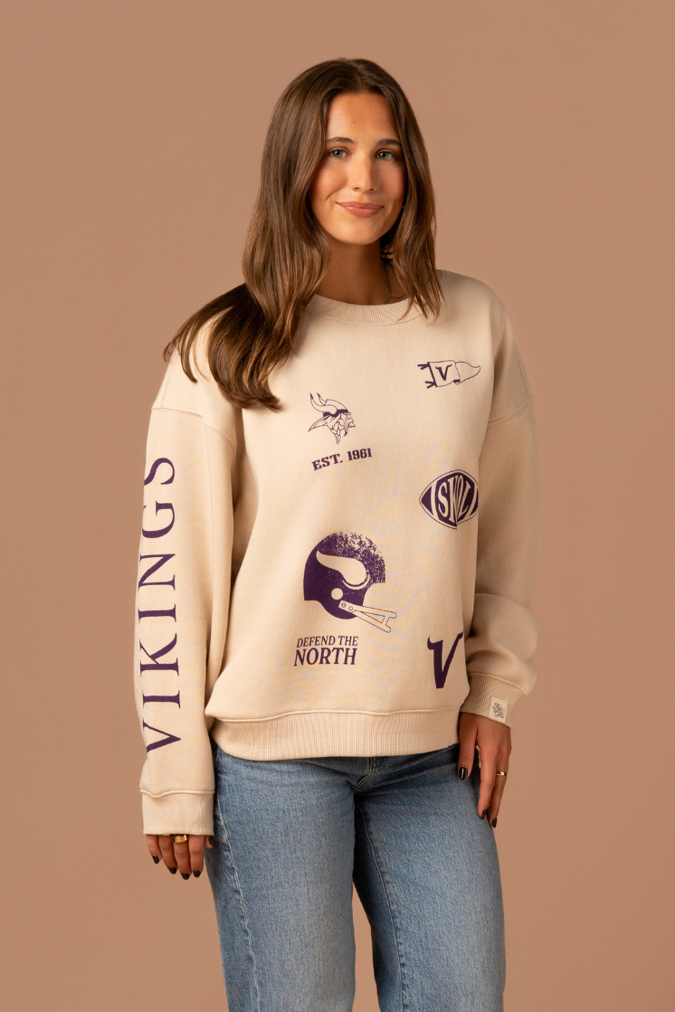 Woman wearing a tan crew neck sweatshirt with five, small purple Minnesota Vikings graphics scattered around the front with purple 'Vikings' text down the arm