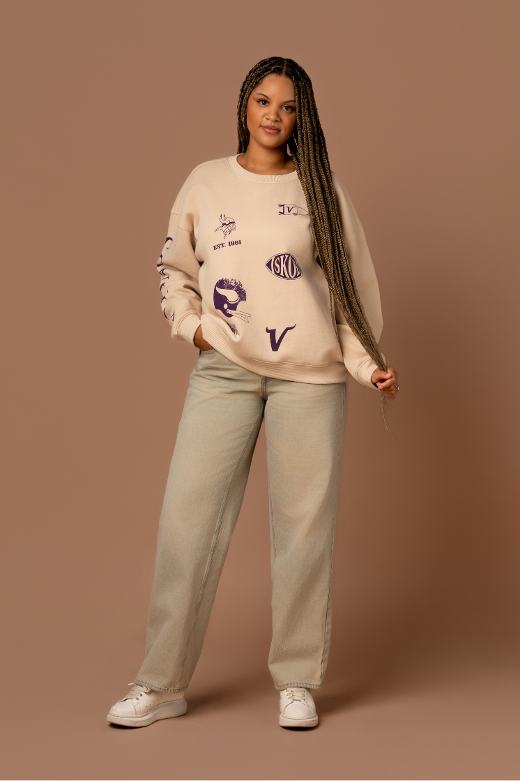 Woman wearing a tan crew neck sweatshirt with five, small purple Minnesota Vikings graphics scattered around the front with purple 'Vikings' text down the arm
